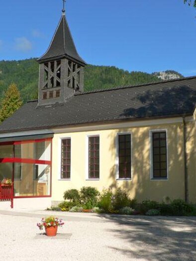 A beautiful chapel with a tall tower and large windows. It is located in a green environment, surrounded by mountains and trees. | © Evangelische Kirche Stainach