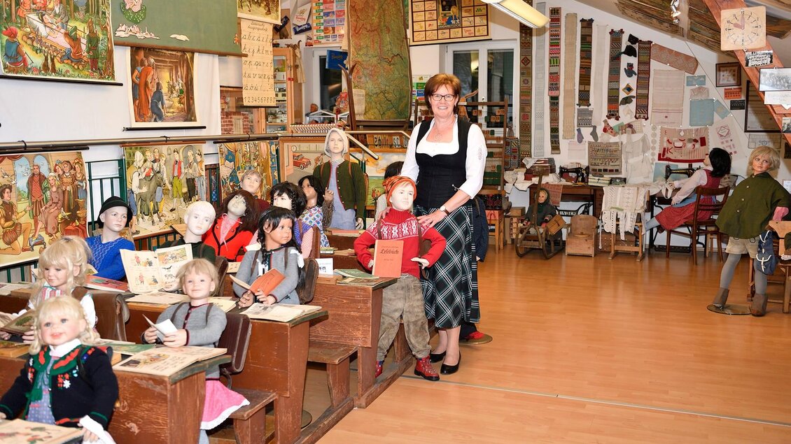 Ein Klassenzimmer mit vielen alten Puppen, die als Schüler an Tischen sitzen. Eine Lehrerin steht lächelnd in der Mitte und umgeben von historischen Bildern und Lernmaterialien. | © Eva Kohlbacher