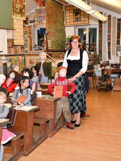 A classroom with many old dolls sitting at desks as students. A teacher stands smiling in the center, surrounded by historical pictures and learning materials. | © Eva Kohlbacher