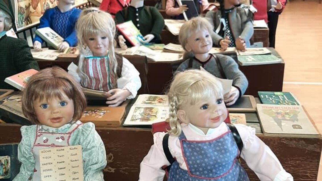 Ein Klassenzimmer mit zahlreichen Puppen, die an Schreibtischen sitzen und Bücher auf den Tisch haben. Die Wand ist mit bunten Bildern dekoriert. | © Eva Kohlbacher