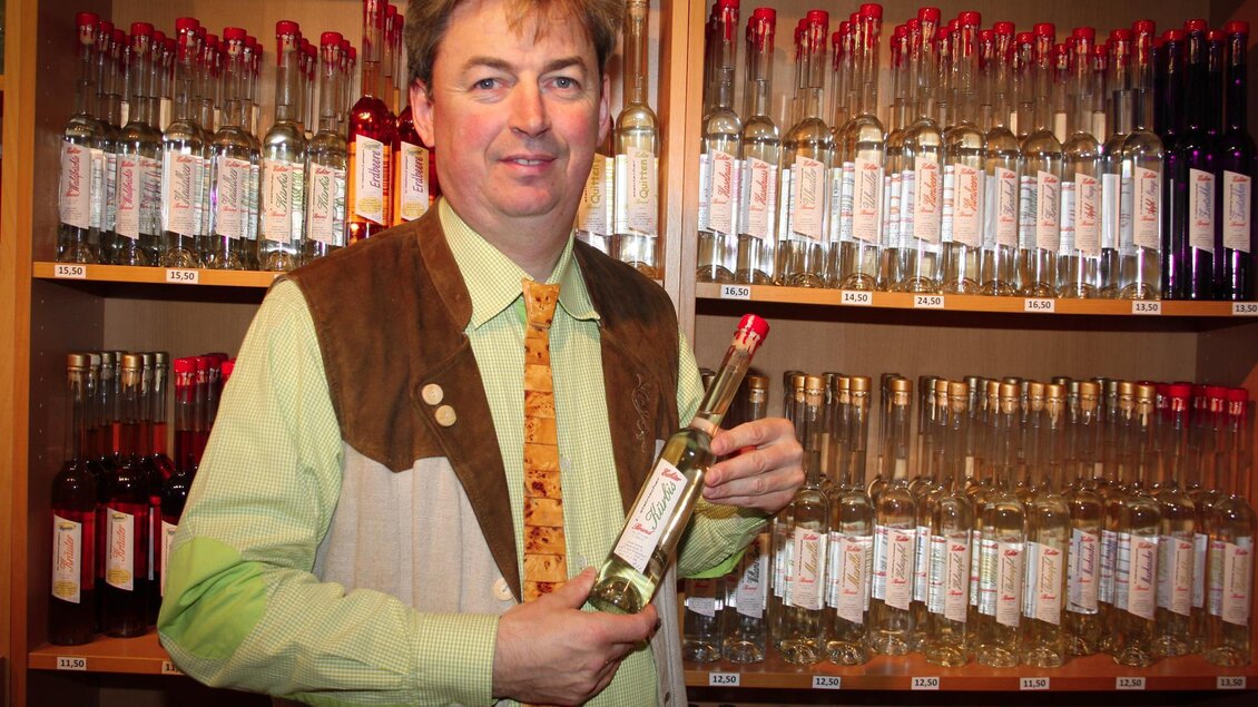 Ein Mann in traditioneller Kleidung hält eine Flasche in der Hand. Im Hintergrund stehen viele Flaschen auf Regalen. | © Lederer