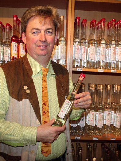 Ein Mann in traditioneller Kleidung hält eine Flasche in der Hand. Im Hintergrund stehen viele Flaschen auf Regalen. | © Lederer