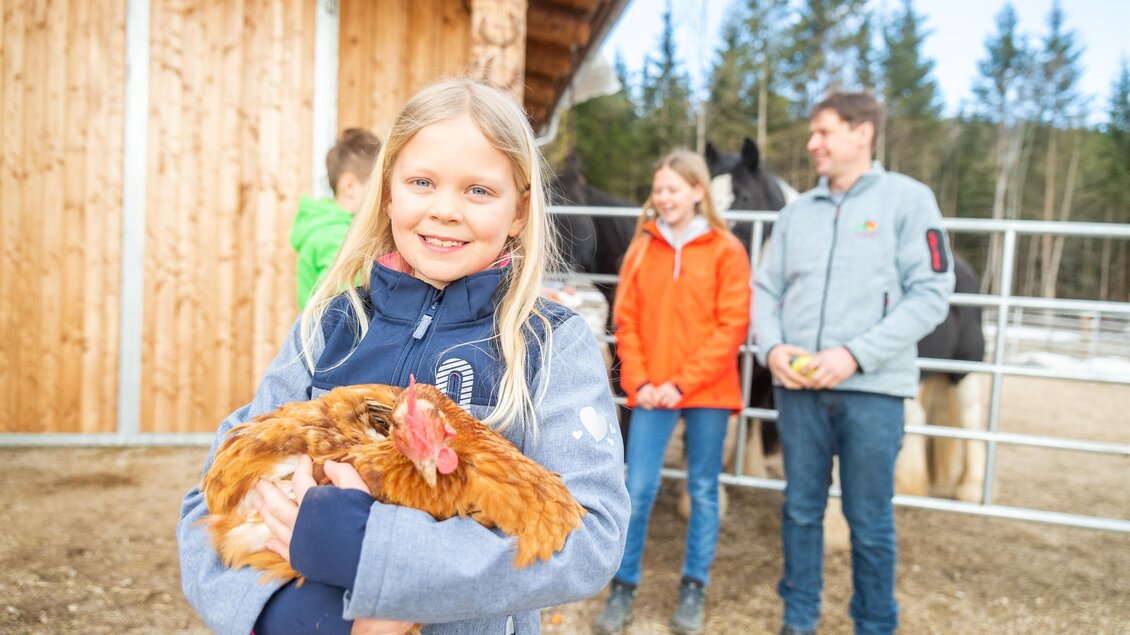 Ein Mädchen hält ein Huhn in den Armen und lächelt. Im Hintergrund stehen weitere Kinder und ein Mann neben Pferden. | © LK - Danner
