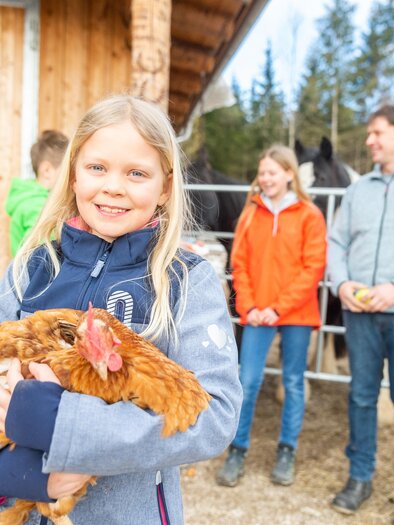 Ein Mädchen hält ein Huhn in den Armen und lächelt. Im Hintergrund stehen weitere Kinder und ein Mann neben Pferden. | © LK - Danner