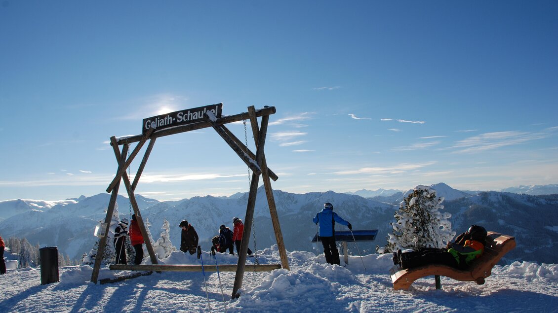 Ein schneebedeckter Berg mit einer Holzschaukel und mehreren Personen im Hintergrund. Der Himmel ist klar und strahlend blau. | © Reiteralm Bergbahnen