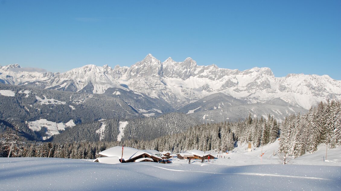 Eine verschneite Berglandschaft mit hohen Gipfeln und klarem Himmel. Im Vordergrund liegen sanfte Schneehügel und einige Berghütten. | © Reiteralm Bergbahnen