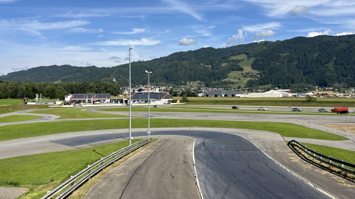 Eine Landschaft mit einem Rundkurs und einer grünen Wiese. Im Hintergrund sind Berge und einige Gebäude sichtbar. | © Peter Hopf