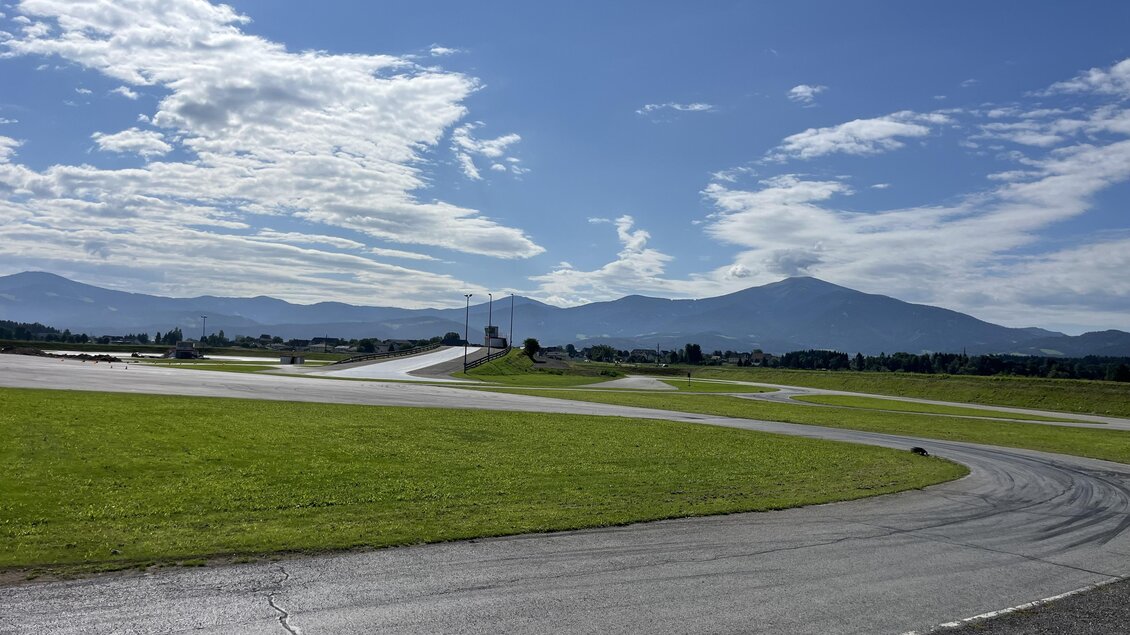Eine Rennstrecke umgeben von grünen Wiesen und Bergen im Hintergrund. Der Himmel ist klar mit einigen Wolken. | © Peter Hopf