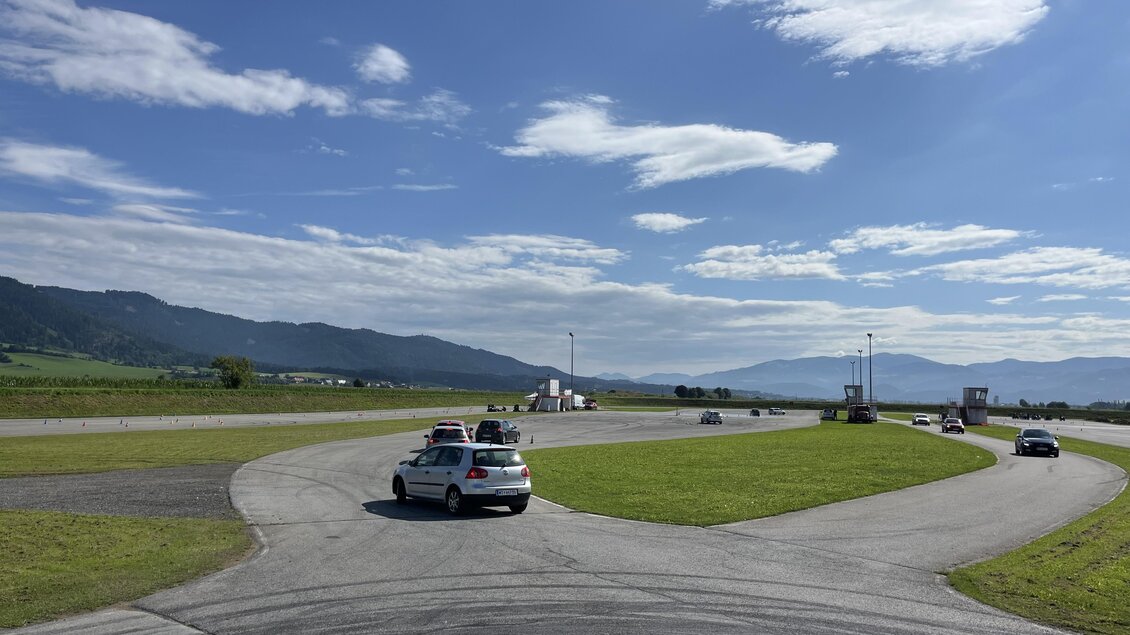 Eine Rennstrecke mit mehreren Autos, die fahren. Im Hintergrund sind Berge und ein blauer Himmel mit Wolken zu sehen. | © Peter Hopf