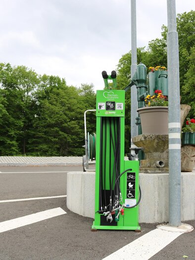 A green charging station for electric vehicles is located in a parking lot. In the background, there are trees and a road visible. | © TVB Thermen- & Vulkanland