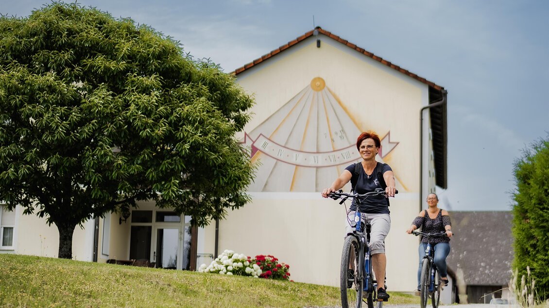 Zwei Personen fahren mit Fahrrädern auf einem schmalen Weg. Im Hintergrund ist ein Gebäude mit einer bunten Wandgestaltung und viel Grün zu sehen. | © Teuschler_Mogg