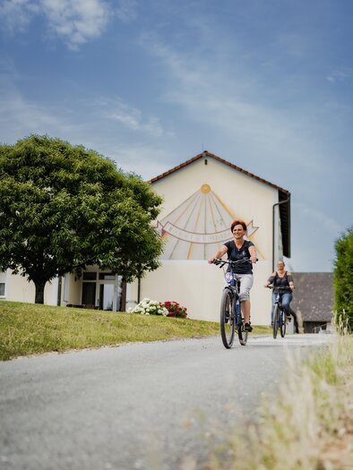 Two people are riding bicycles on a narrow path. In the background, there is a building with colorful wall design and a lot of greenery. | © Teuschler_Mogg