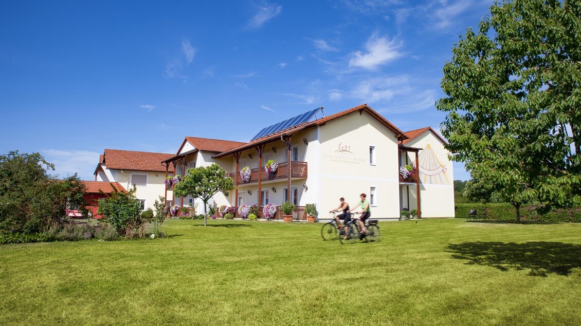Ein modernes Gebäude mit hübschen Balkonen und grünen Pflanzen umgeben. Im Vordergrund fahren zwei Personen mit dem Fahrrad über eine grüne Wiese. | © Fotospass