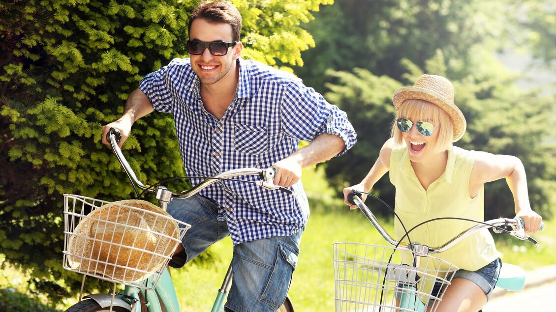 Ein fröhliches Paar fährt gemeinsam Fahrrad in einer sonnigen Umgebung. Beide tragen Sonnenbrillen und genießen den Moment. | © fotolia