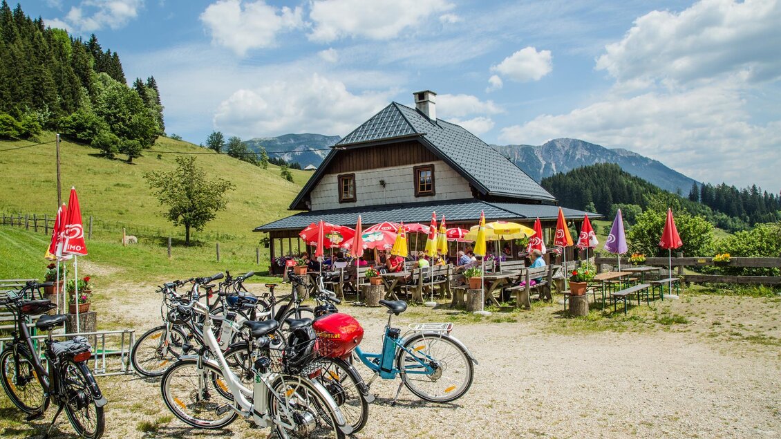Ein gemütliches Restaurant in der Natur mit Terrassenplatz und bunten Sonnenschirmen. Vor dem Gebäude stehen Fahrräder auf einem Schotterweg. | © Naturpark Mürzer Oberland
