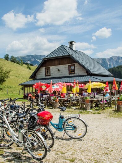 A cozy restaurant in nature with a terrace seating area and colorful sun umbrellas. Bicycles are parked on a gravel path in front of the building. | © Naturpark Mürzer Oberland
