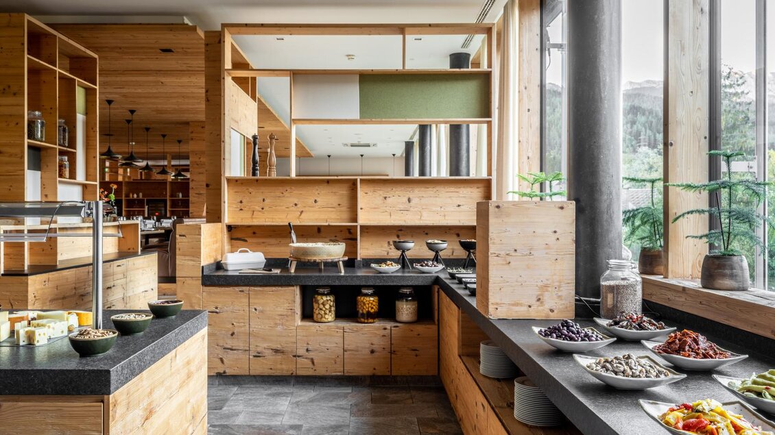 Eine moderne Küche mit Holzregalen und einer Vielzahl an frischen Zutaten. Der Raum ist hell und einladend, mit großen Fenstern und stilvollen Details. | © Falkensteiner Hotel Schladming/ Fotograf Dragan Dokmanovic