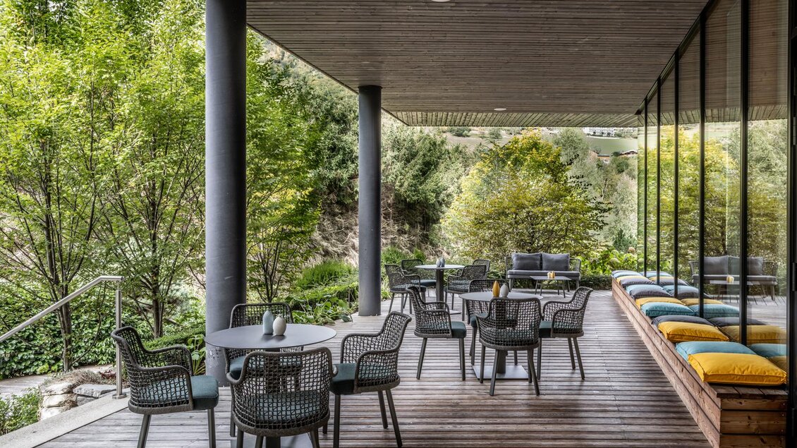 Ein stilvolles Außensitzbereich mit modernen Möbeln und viel Grün. Der Holzfußboden und die Glaswände bieten einen gemütlichen Ausblick auf die Natur. | © Falkensteiner Hotel Schladming/ Fotograf Dragan Dokmanovic