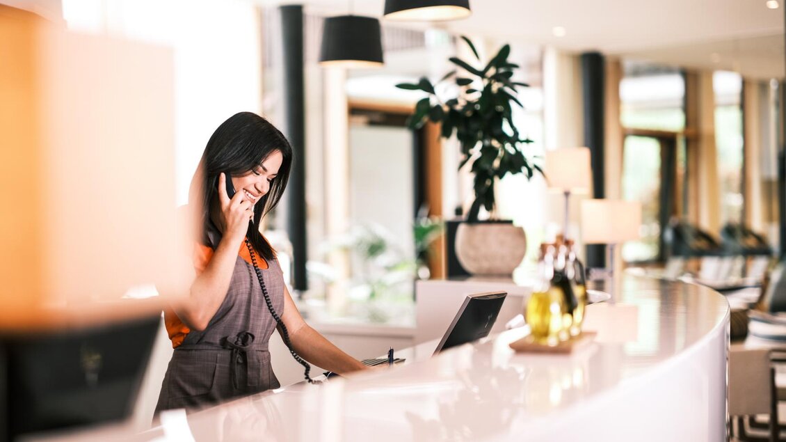 Eine Empfangsdame spricht am Telefon in einem modernen Hotel. Der Raum ist hell und freundlich mit Pflanzen und gemütlichen Sitzgelegenheiten. | © Falkensteiner Hotel Schladming/ Fotograf Dragan Dokmanovic