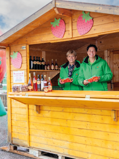 Ein kleiner Holzstand verkauft frische Erdbeeren und Getränke. Im Hintergrund steht eine große aufblasbare Erdbeere. | © Bauers Beeradies