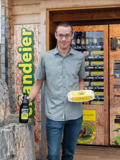 A man is standing in front of a vending machine for food, holding a pack of eggs and a bottle of oil. Next to him is a wooden sculpture of a chicken. | © Familie Passath