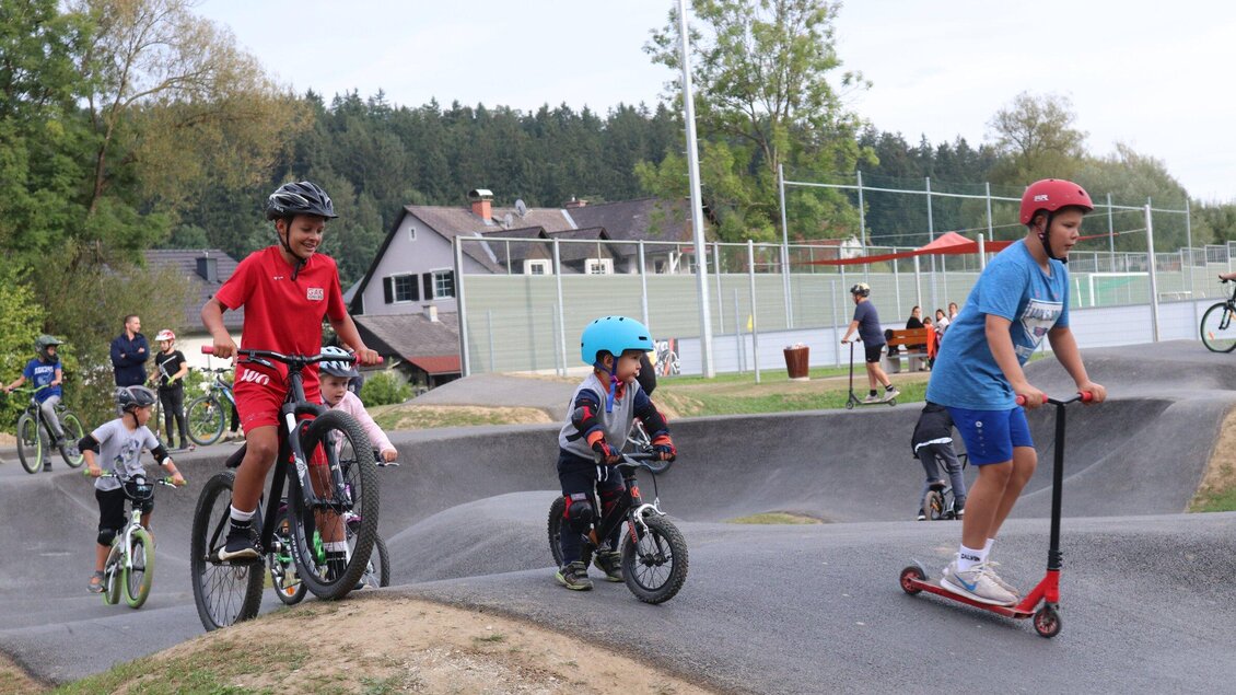 Kinder fahren mit Fahrrädern und Rollern auf einem Pumptrack. Im Hintergrund sind Bäume und ein Sportplatz sichtbar. | © Gemeinde Ligist-Cescutti Robert