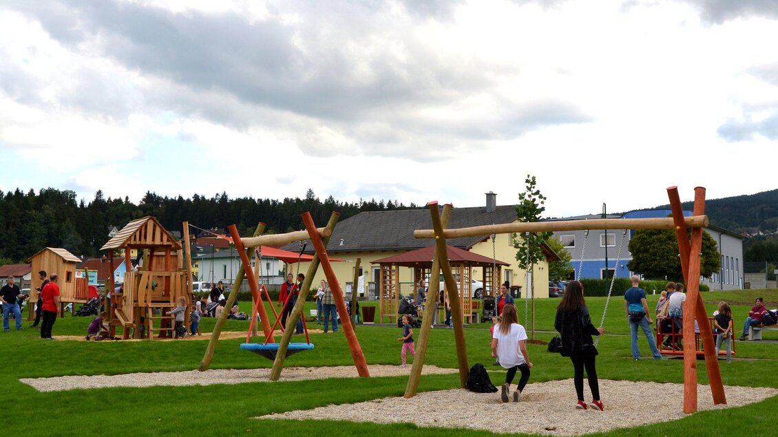 Ein Spielplatz mit modernen Schaukeln und einem Holzspielhaus. Viele Kinder und Erwachsene genießen die Zeit im Freien. | © Gemeinde Ligist-Kremser Gottfried