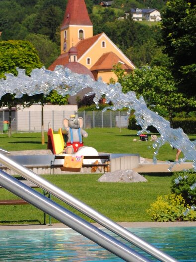 A beautiful garden with a pool and a water feature. In the background, there are green trees and a small church. | © Marktgemeinde Ligist
