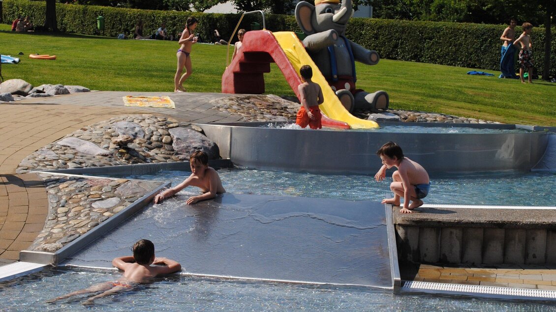 Kinder planschen in einem kleinen Schwimmbecken mit einem Wasserrutsch. Im Hintergrund gibt es eine gepflegte Rasenfläche und Freizeitaktivitäten. | © Marktgemeinde Ligist