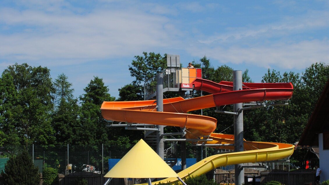 Eine Wasserbahn mit bunten Rutschen in einem Freibad. Im Hintergrund sind Bäume und ein blauer Himmel zu sehen. | © Marktgemeinde Ligist
