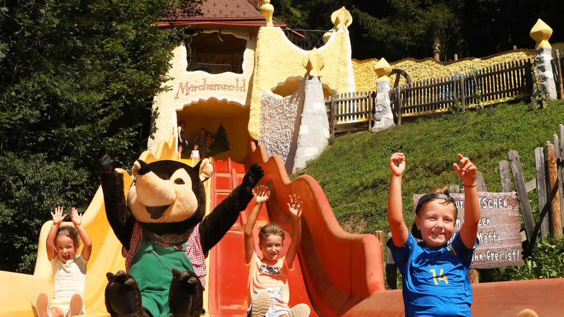 Ein fröhlicher Wasserrutschenpark mit Kindern, die Spaß haben. Ein Maskottchen sitzt neben ihnen und alle haben die Hände in die Luft gehoben. | © Märchenwald