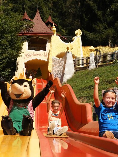 A cheerful water slide park with children having fun. A mascot is sitting next to them and everyone has their hands raised in the air. | © Märchenwald