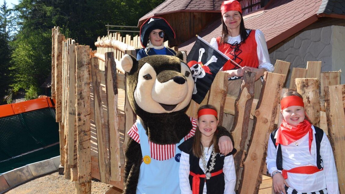 Eine Gruppe von Verkörperungen in Piratenkostümen steht an einer Holzkonstruktion. Ein großer Bärenmaskottchen lächelt und posiert mit zwei Kindern und einer Erwachsenen. | © Märchenwald