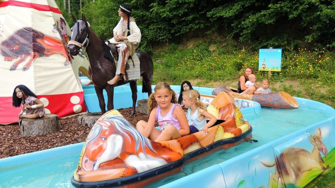 Ein Kinderbootfahren in einem Wasserbecken, umgeben von einer Figur auf einem Pferd und einem Tipi. Im Hintergrund sind fröhliche Kinder, die Spaß haben. | © Märchenwald