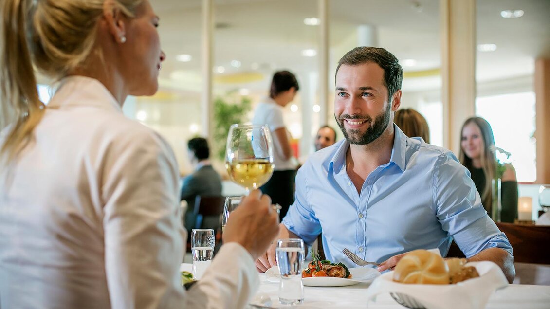 Ein stilvolles Restaurant mit zwei Personen, die einander im Gespräch an einem Tisch gegenüber sitzen. Der Mann lächelt und hält ein Weinglas in der Hand. | © H2O Hotel-Therme-Resort