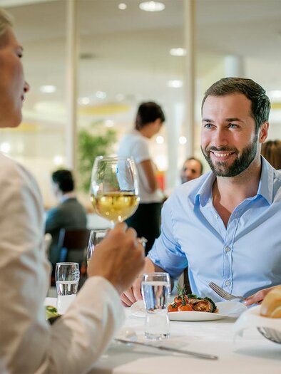 A stylish restaurant with two people sitting across from each other at a table engaged in conversation. The man is smiling and holding a wine glass in his hand. | © H2O Hotel-Therme-Resort