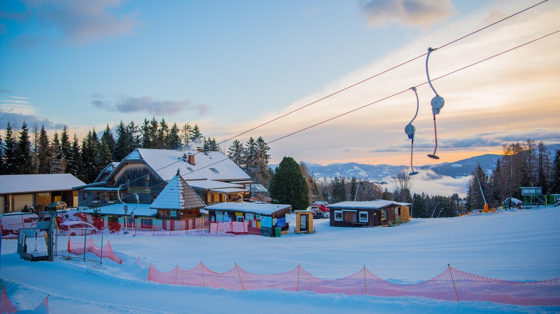 Eine verschneite Landschaft mit einem Skilift und einer Berghütte. Der Himmel ist klar und zeigt sanfte Farben bei Sonnenuntergang. | © Anita Fössl