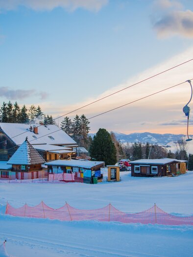 A snowy landscape with a ski lift and a mountain hut. The sky is clear and shows gentle colors at sunset. | © Anita Fössl