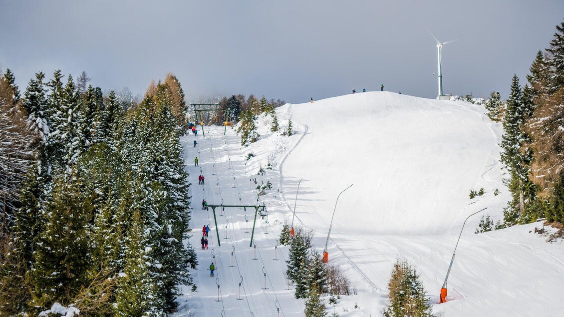 Eine schneebedeckte Skipiste mit Lifts, umgeben von hohen Tannenbäumen. Im Hintergrund ist ein sanfter Hang zu sehen, ideal für Wintersport. | © Anita Fössl
