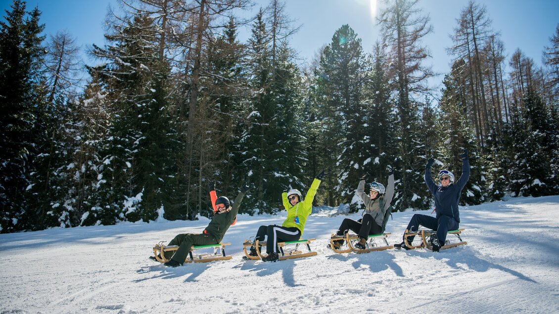 Eine Gruppe von vier Personen sitzt auf Schlitten im Schnee und hebt die Hände in die Luft. Um sie herum sind schneebedeckte Bäume und ein strahlend blauer Himmel. | © Anita Fössl