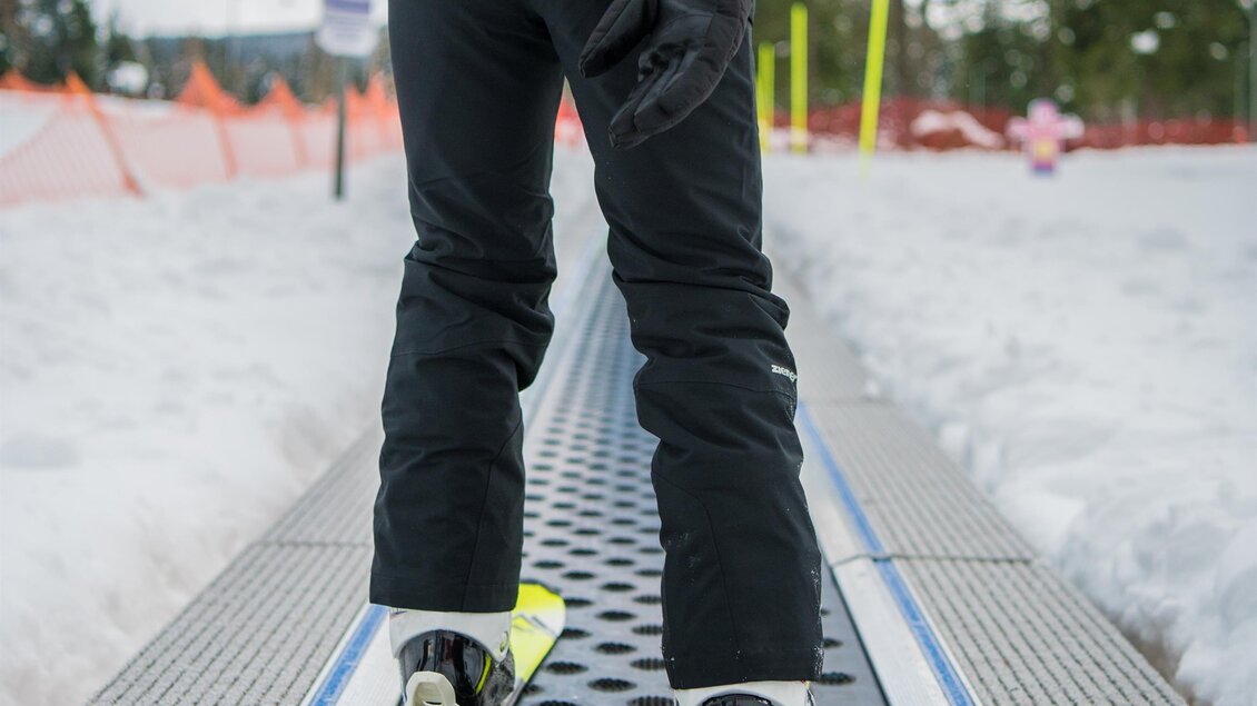 Eine Person auf Skiern steht auf einem Förderband im Schnee. Im Hintergrund sind Bäume und ein blauer Himmel zu sehen. | © Anita Fössl