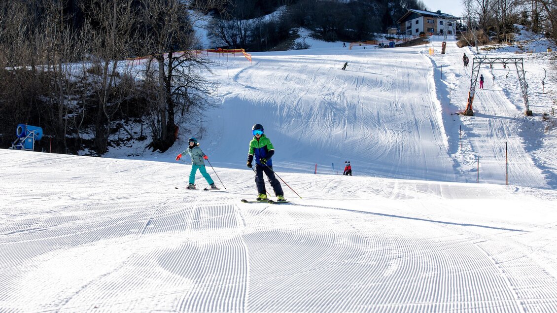 Zwei Skifahrer auf einer schneebedeckten Piste in den Bergen. Im Hintergrund sind Bäume und eine Skiliftstation zu sehen. | © Tourismusverband Murau