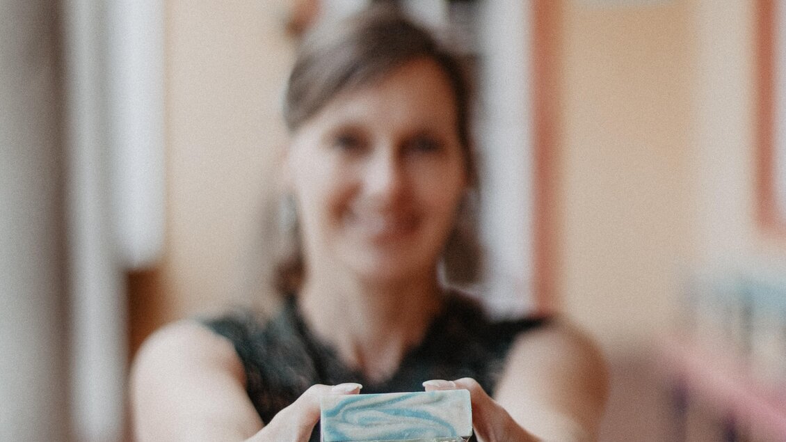 Eine Frau hält eine stilvoll verpackte Seife in den Händen. Der Hintergrund zeigt eine hellfarbige Wand und eine Tür. | © Ute Oberzauchner 