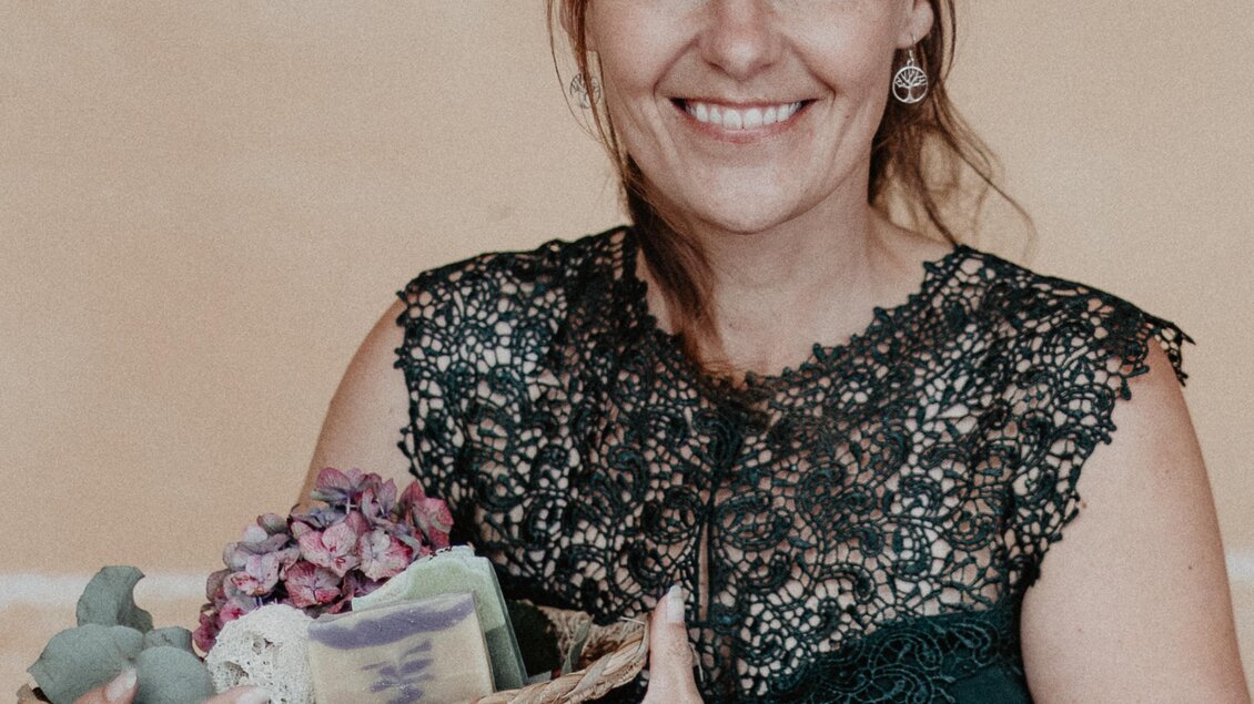 Eine lächelnde Frau hält einen Korb mit getrockneten Blumen. Sie trägt ein schickes schwarzes Oberteil mit Spitze. | © Ute Oberzauchner 