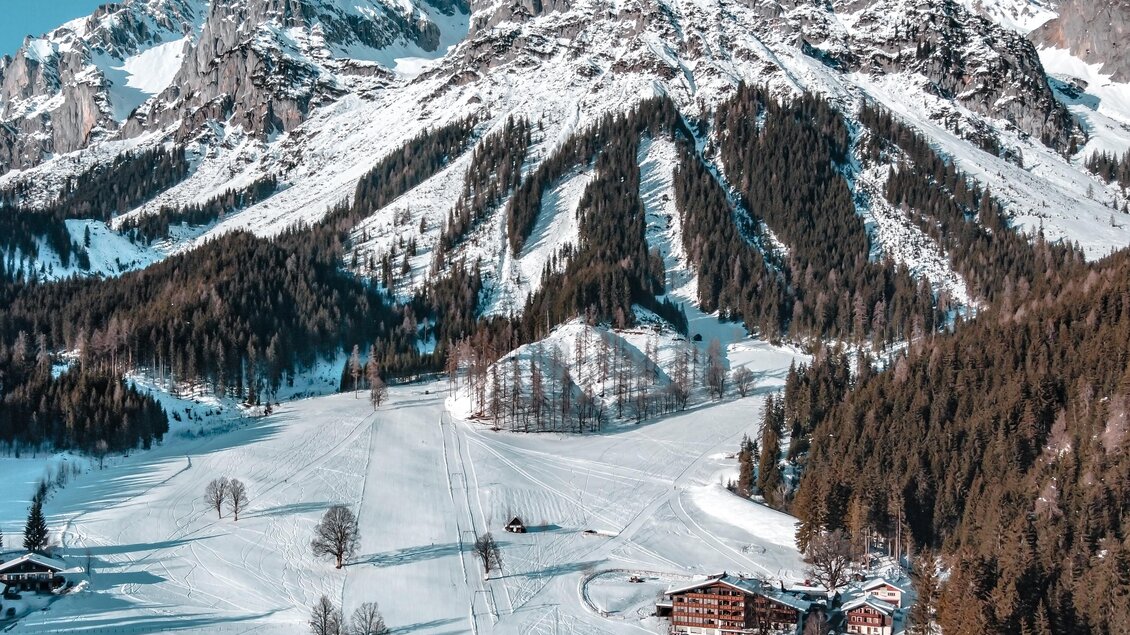 Eine verschneite Berglandschaft mit hohen Gipfeln und dichten Wäldern. Im Vordergrund sind einige Hütten und eine Skipiste sichtbar. | © Anna Ullrich