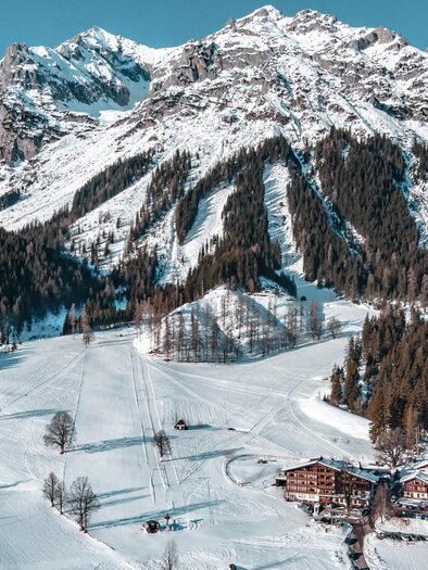 A snowy mountain landscape with high peaks and dense forests. In the foreground, some huts and a ski slope are visible. | © Anna Ullrich