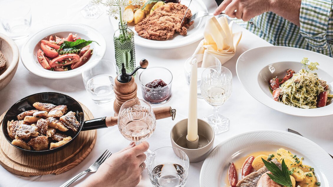 Ein gedeckter Tisch mit verschiedenen Speisen, darunter Fleisch und frische Salate. Dazu gehören Gläser mit Wasser und Wein sowie Kerzenlicht für eine angenehme Atmosphäre. | © Anna Ullrich