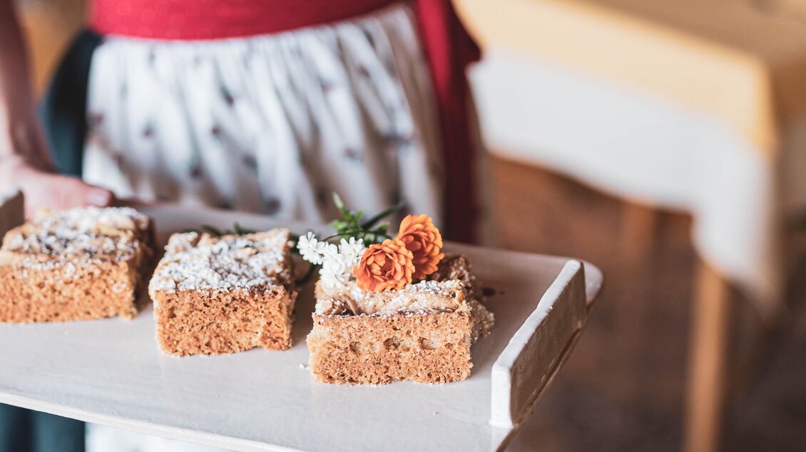 Ein Tablett mit leckeren Kuchenstücken, dekoriert mit Blumen. Im Hintergrund ist ein gemütlicher Raum zu sehen. | © Anna Ullrich