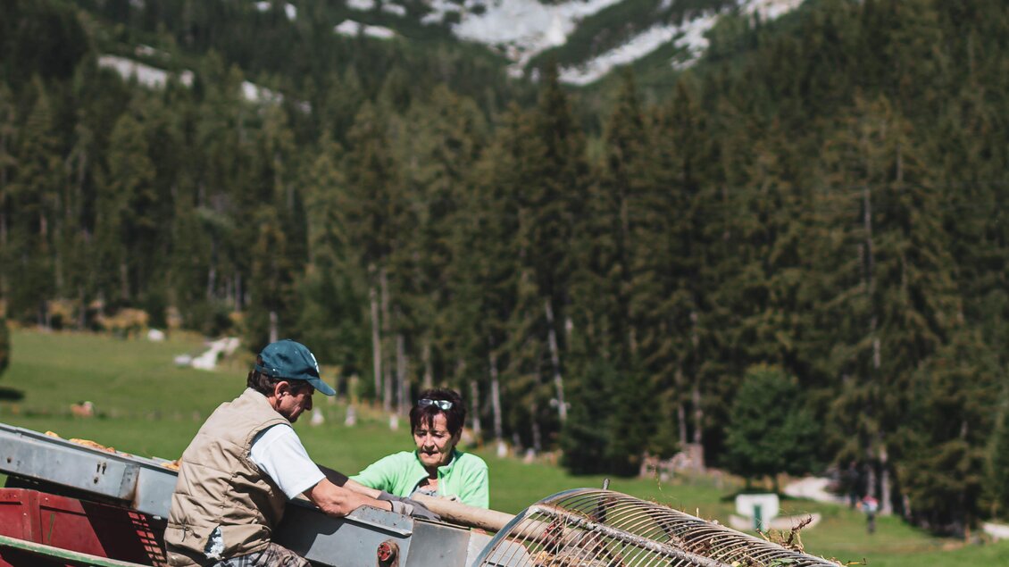 Ein Landwirt und ein Helfer arbeiten mit einer Maschine auf einem Feld in den Bergen. Im Hintergrund sind majestätische Berge und Wälder zu sehen. | © Anna Ullrich