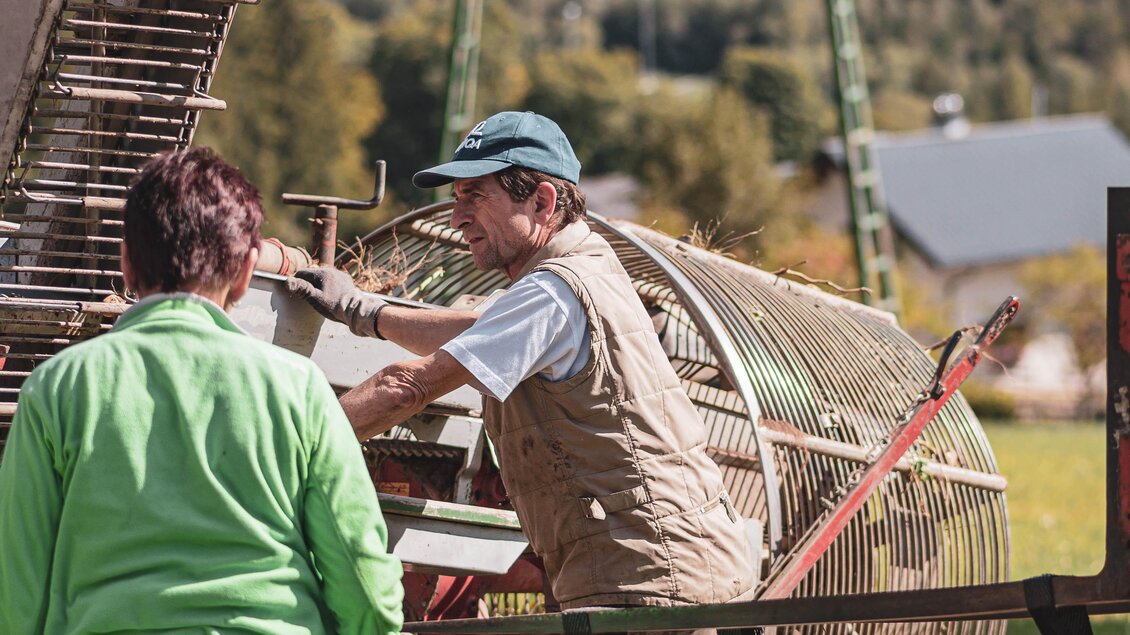 Ein Mann und eine Frau arbeiten zusammen auf einem Feld, während eine Maschine zur Ernte genutzt wird. Im Hintergrund sind Bäume und ein Gebäude zu sehen. | © Anna Ullrich